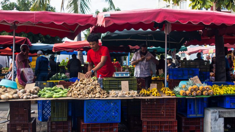 Un forain (bazardier) range des légumes derrière son étal sur lequel on trouve plusieurs variétés de fruits et légumes (des patates douces, des morceaux de gingembre, des bananes, des ananas